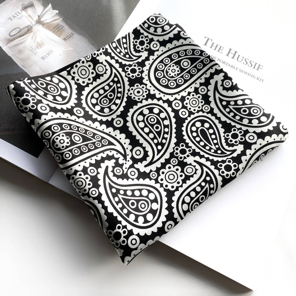 Black silk scarf with white paisley pattern folded into a square shape on a white background on a magazine.