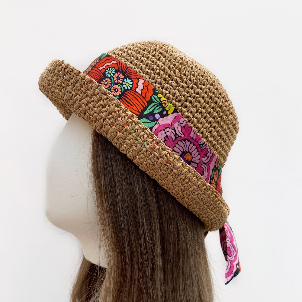 Liberty silk skinny scarf in Begonia Garden floral print tied on a brown straw hat on a mannequin head against a white background
