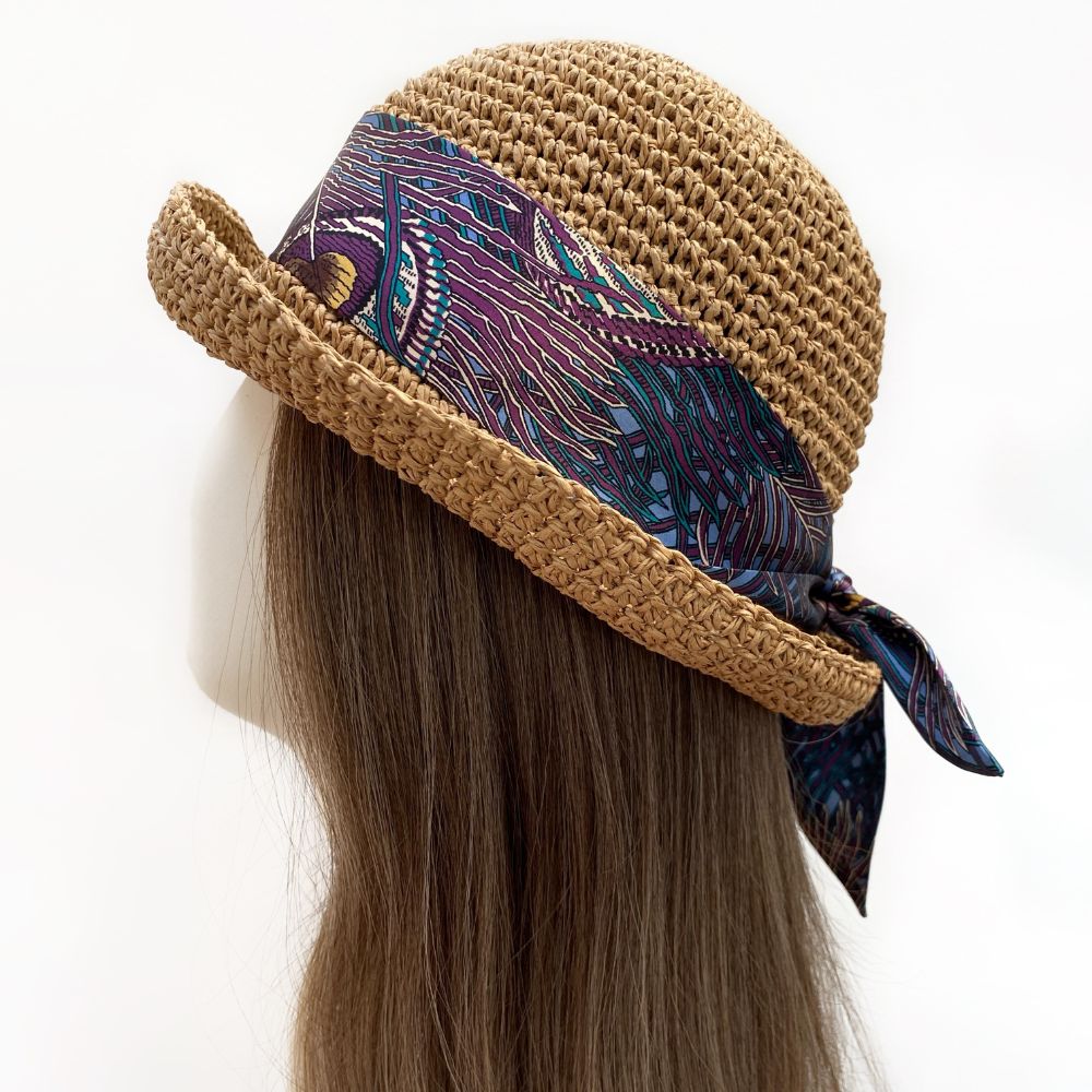 Brown straw hat with a Liberty silk skinny scarf featuring the Royal Hera peacock print on a mannequin head against a white background