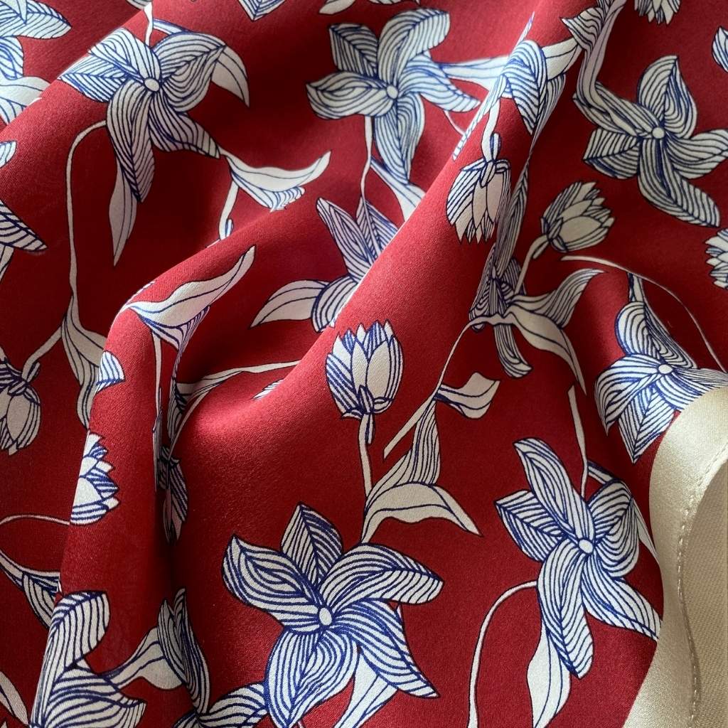 Close-up of a red silk scarf with a blue and white sketched floral pattern, featuring a cream-colored border.