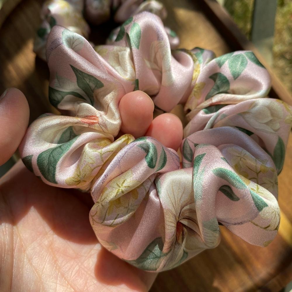 A close-up of a liberty silk satin hair scrunchie with a light pink base and a green and yellow floral print, held in a person's hand.
