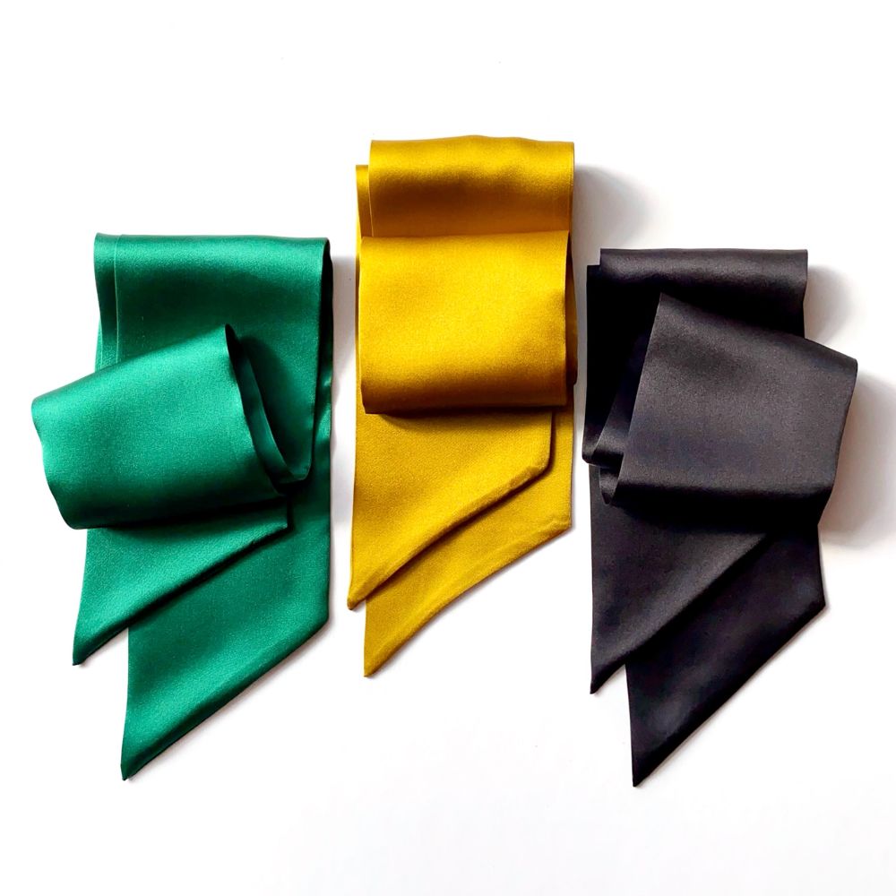 Three folded mulberry silk satin neck scarves in emerald green, golden yellow, and charcoal black, arranged side-by-side on a plain white background.