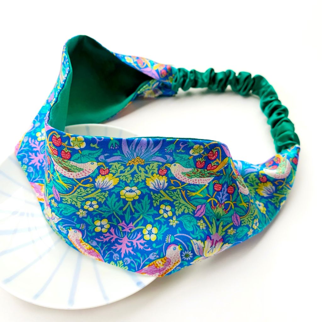 A  Liberty London mulberry silk satin headband with a vibrant william morris strawberry thief pattern, featuring a dark green elastic band, displayed on a white plate against a white background.