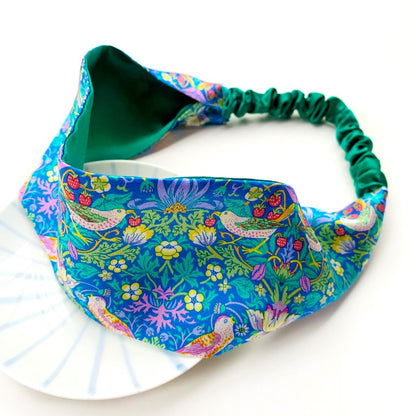 A  Liberty London mulberry silk satin headband with a vibrant william morris strawberry thief pattern, featuring a dark green elastic band, displayed on a white plate against a white background.