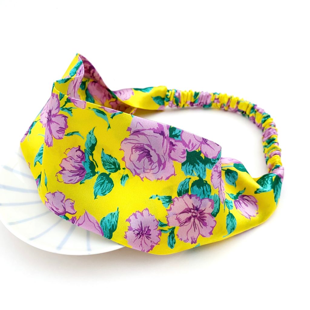 A bright yellow  Liberty London mulberry silk satin headband with a pink and green rose floral print, displayed on a white surface.