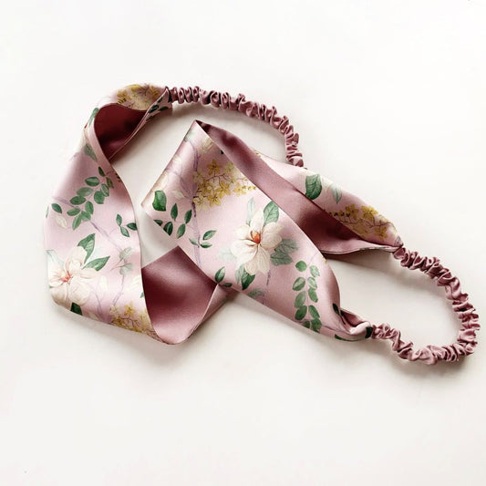 A pink Liberty London silk headband with a floral print in shades of dusty pink, white, and green, paired with matching dusty rose elastic back, displayed on a clean white background.