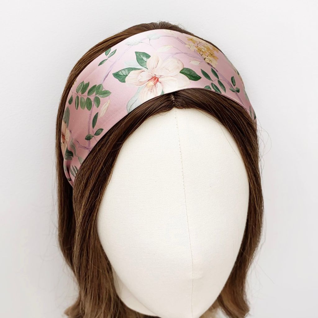 A wide pink  Liberty London mulberry silk satin headband with a floral print, displayed on a mannequin head with brown hair against a plain white background.