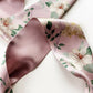 A close-up of a  Liberty London mulberry silk satin headband with a dusty pink base, decorated with white magnolia flowers, green leaves, and small yellow blossoms.