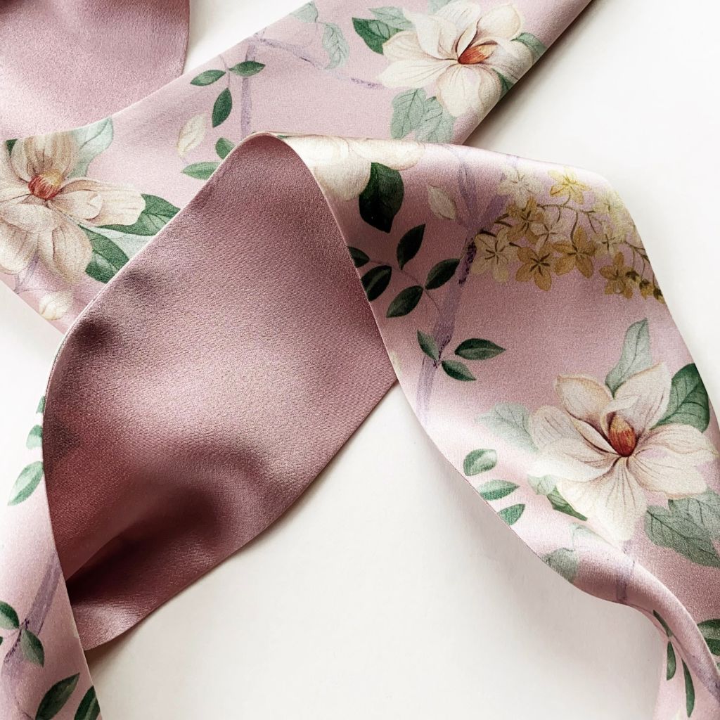 A close-up of a  Liberty London mulberry silk satin headband with a dusty pink base, decorated with white magnolia flowers, green leaves, and small yellow blossoms.