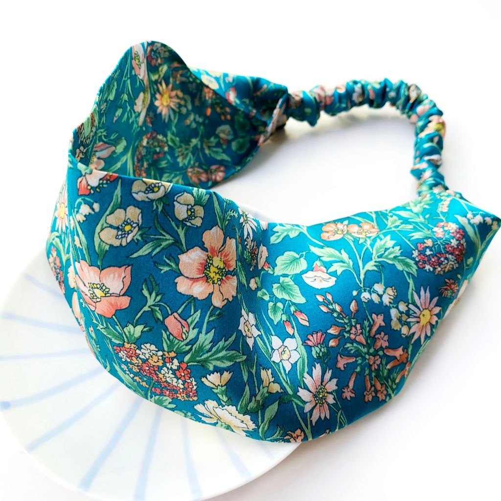 A wide  Liberty London mulberry silk satin headband with an elastic back, featuring a vibrant teal blue background with a dense multicolor floral print of pink, white, and yellow flowers with green leaves. The headband is styled on a white plate against a clean white background.