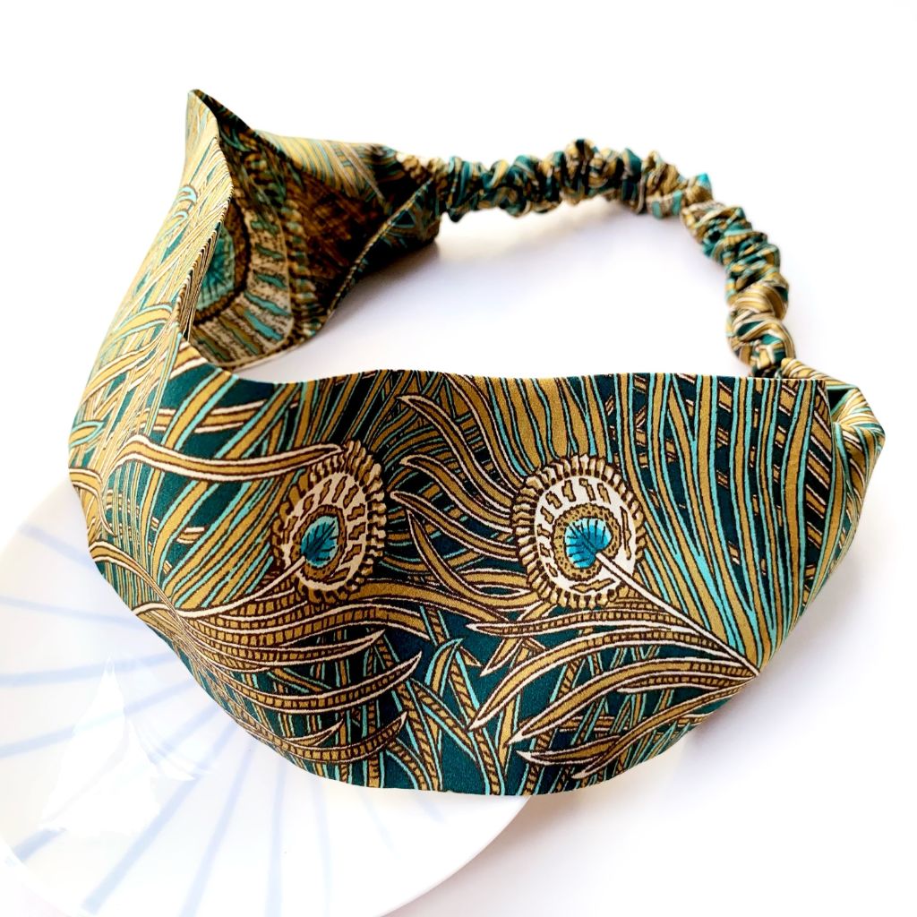 A  Liberty London mulberry silk satin headband featuring a detailed peacock feather pattern in teal green, gold, and brown on a white background.
