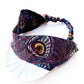 A  Liberty London mulberry silk satin headband with a peacock feather pattern in purple, teal, black, and gold, displayed on a white plate against a white background.