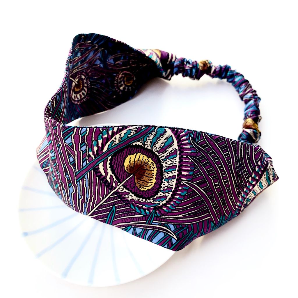 A  Liberty London mulberry silk satin headband with a peacock feather pattern in purple, teal, black, and gold, displayed on a white plate against a white background.