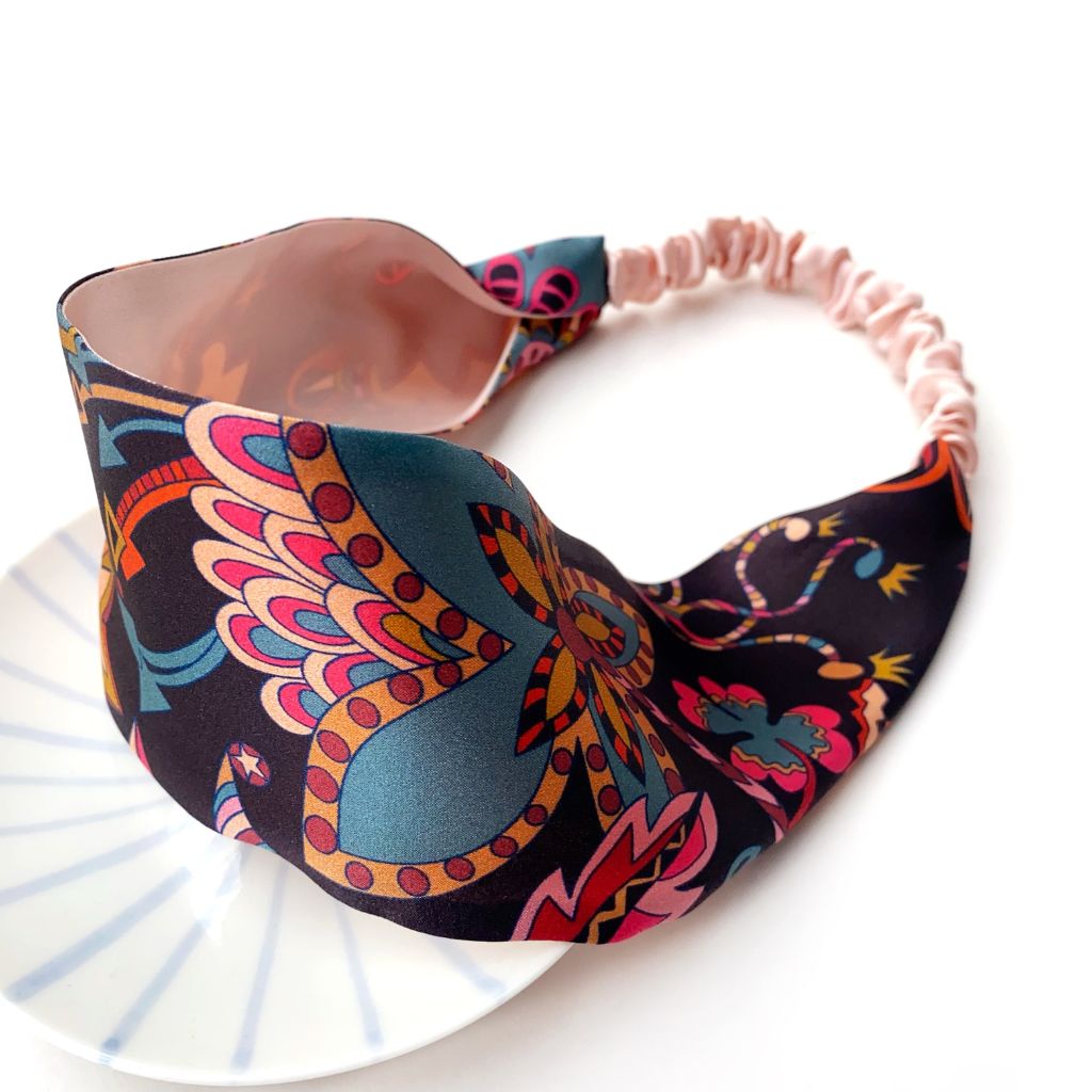 A  Liberty London mulberry silk satin headband with a colorful pattern on a black background, featuring a light pink elastic back, displayed on a white plate against a white background.