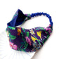 A  Liberty London mulberry silk satin headband with a navy blue silk lining and a colorful floral print, featuring a blue elastic band, displayed on a white plate against a white background.