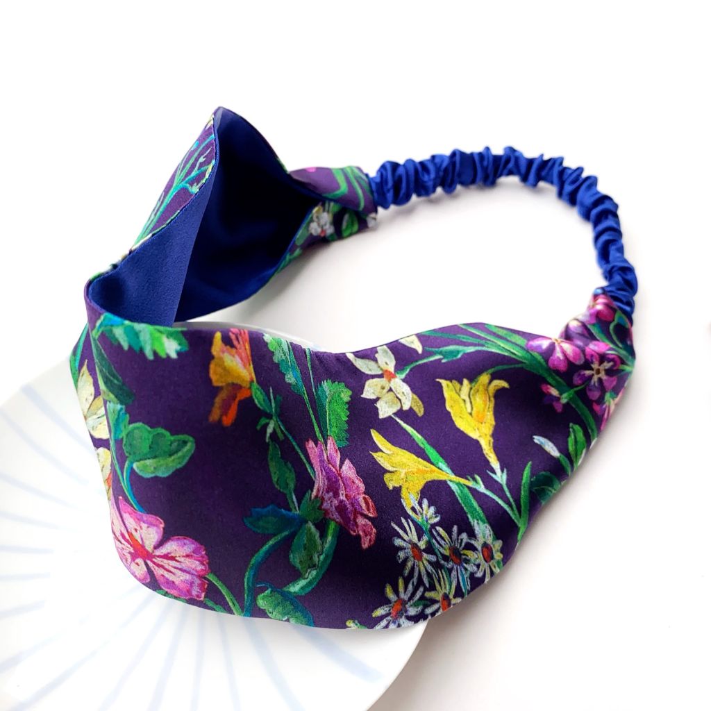 A  Liberty London mulberry silk satin headband with a navy blue silk lining and a colorful floral print, featuring a blue elastic band, displayed on a white plate against a white background.