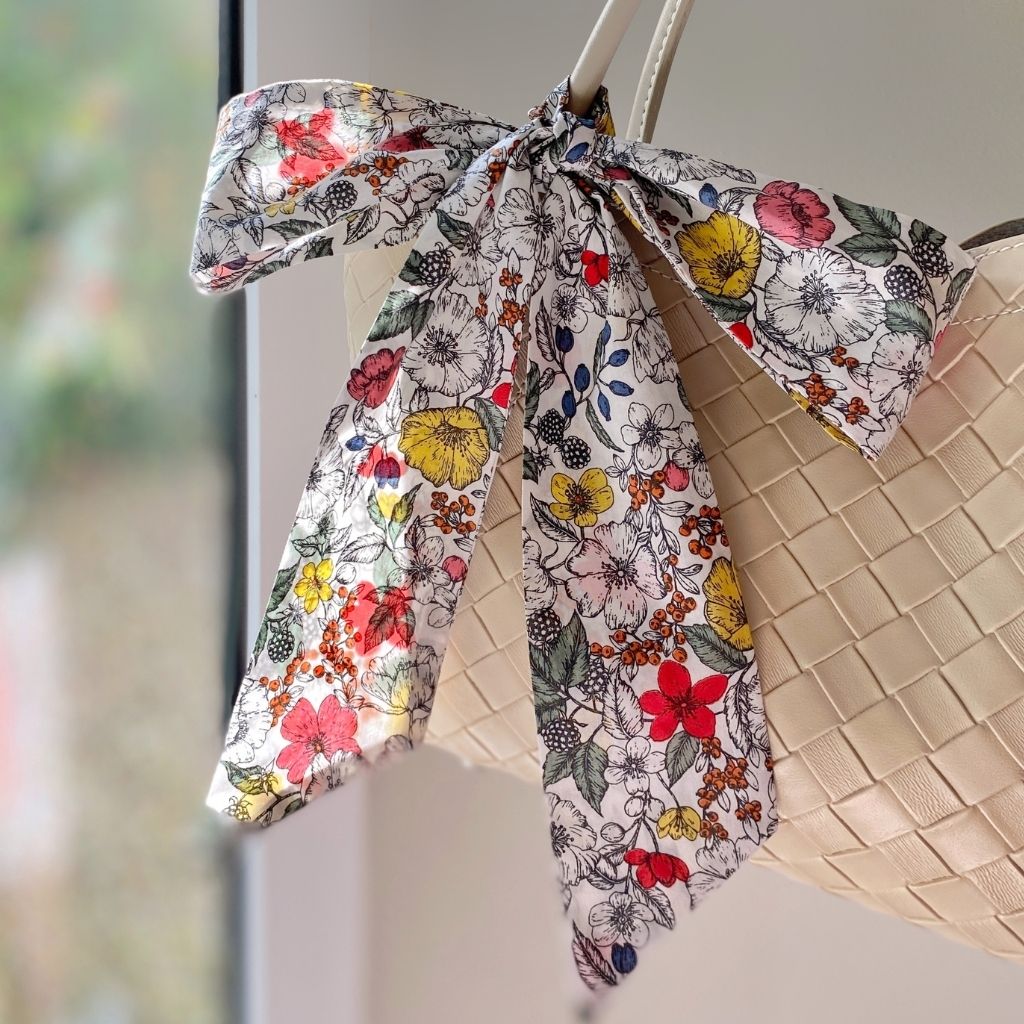 A white and colorful floral print Liberty Tana Lawn cotton skinny scarf tied in a large bow and draped over a woven beige handbag, set against a softly blurred indoor background.