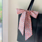 A Liberty Tana Lawn cotton skinny scarf featuring a red floral pattern, tied into a bow on a black handbag, shown hanging indoors next to a window.