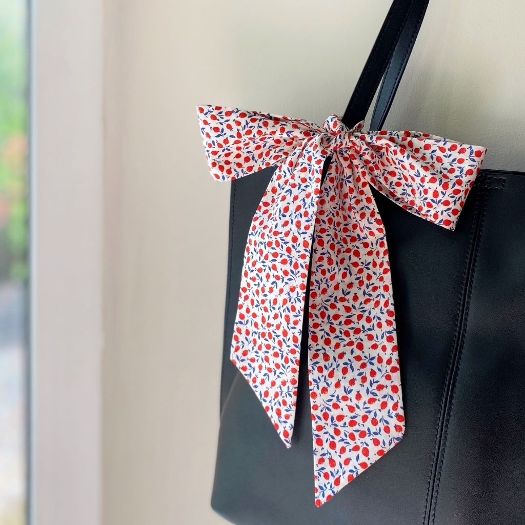 A Liberty Tana Lawn cotton skinny scarf featuring a red floral pattern, tied into a bow on a black handbag, shown hanging indoors next to a window.