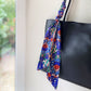 A black leather tote bag with a colorful patterned Liberty Tana Lawn cotton skinny scarf tied to its strap, hanging against a neutral wall.