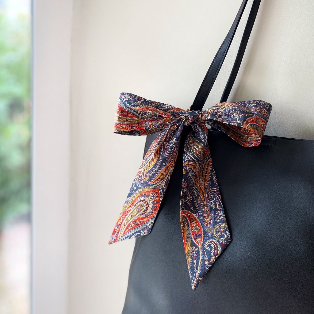 A Liberty Tana Lawn cotton skinny scarf featuring an orange and blue paisley pattern, tied into a bow on a black handbag, shown hanging indoors next to a window.