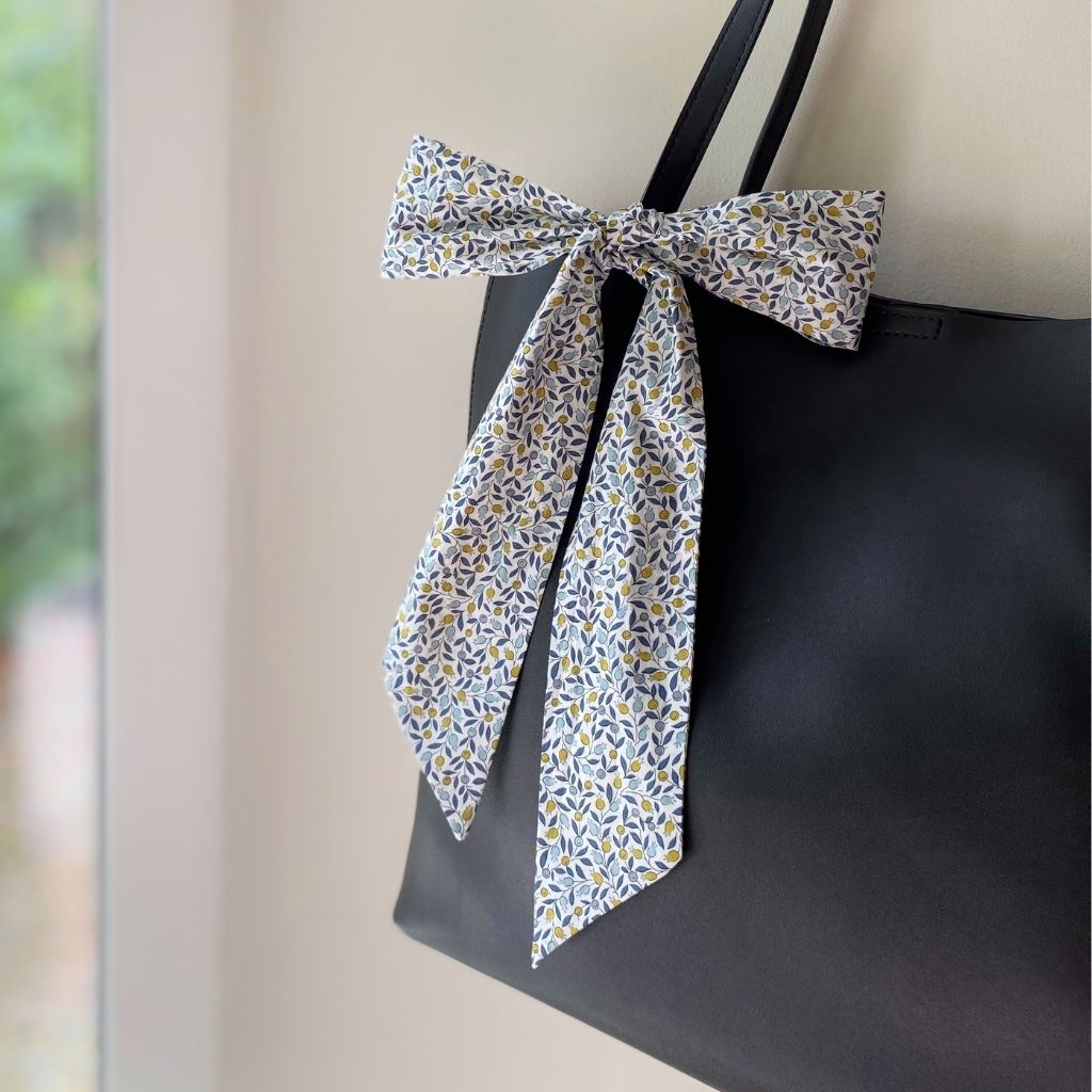 A Liberty Tana Lawn cotton skinny scarf featuring a leafy and small floral pattern, tied into a bow on a black handbag, shown hanging indoors next to a window.