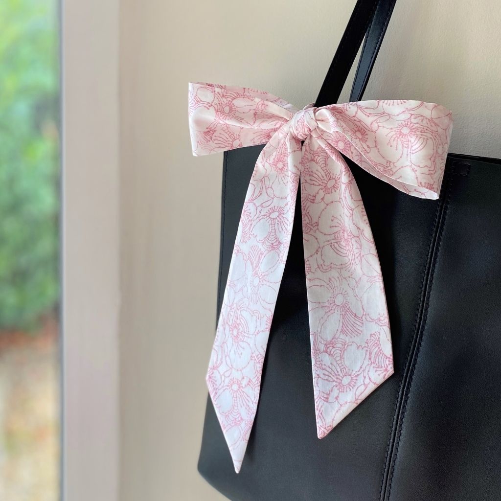 A Liberty Tana Lawn cotton skinny scarf featuring a pink floral pattern, tied into a bow on a black handbag, shown hanging indoors next to a window.