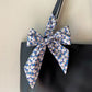 A close-up of a black handbag with a large decorative bow made from a patterned Liberty Tana Lawn cotton skinny scarf. The bow features a white base with a dense floral print in navy blue, light blue, and pink, tied around the black shoulder strap of the bag.