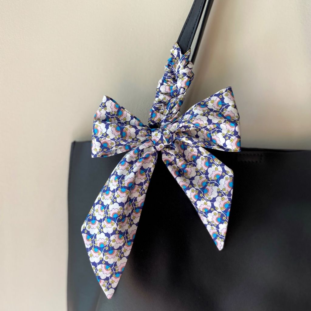 A close-up of a black handbag with a large decorative bow made from a patterned Liberty Tana Lawn cotton skinny scarf. The bow features a white base with a dense floral print in navy blue, light blue, and pink, tied around the black shoulder strap of the bag.