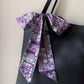 A purple and white Liberty Tana Lawn cotton skinny scarf tied into a large bow on a black tote bag's strap, against a plain light beige wall.