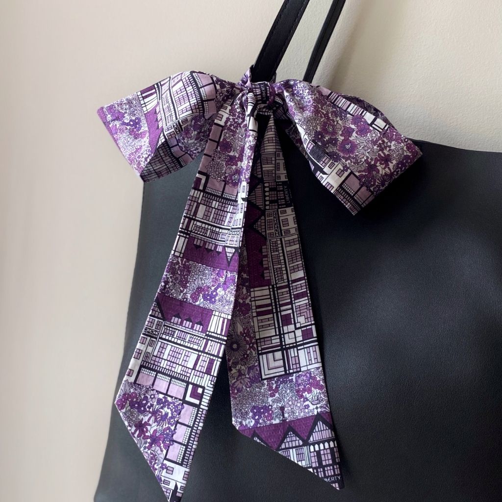 A purple and white Liberty Tana Lawn cotton skinny scarf tied into a large bow on a black tote bag's strap, against a plain light beige wall.