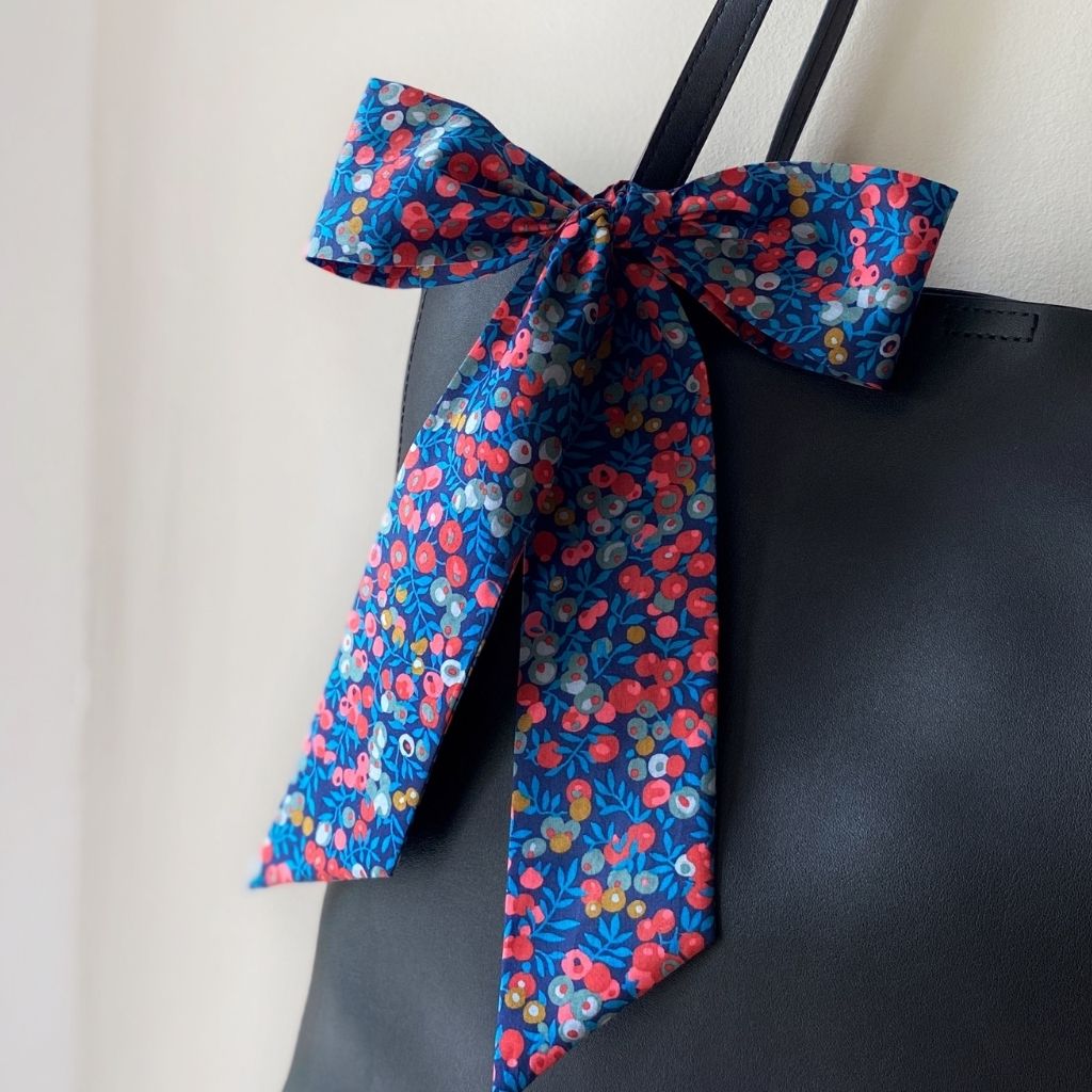 A blue and red berry print Liberty Tana Lawn cotton skinny scarf tied into a large bow on a black tote bag's strap, against a plain light beige wall.