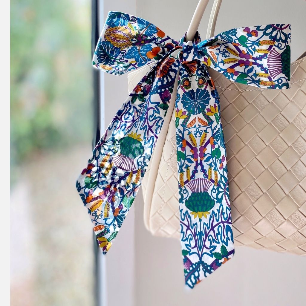 A multicolor patterned Liberty Tana Lawn cotton skinny scarf tied in a large bow and draped over the handle of a cream woven handbag, set against a softly blurred window background.