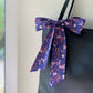An indigo blue Liberty Tana Lawn cotton skinny scarf with a pink and white bunny and floral pattern, tied in a large bow and draped over the shoulder strap of a black tote bag.