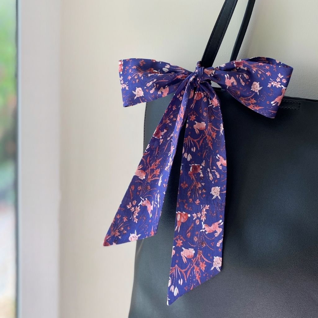 An indigo blue Liberty Tana Lawn cotton skinny scarf with a pink and white bunny and floral pattern, tied in a large bow and draped over the shoulder strap of a black tote bag.
