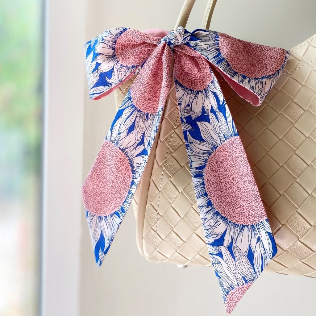 A Liberty Tana Lawn cotton skinny scarf featuring a pink and blue sunflower pattern, tied into a bow on a black handbag, shown hanging indoors next to a window.
