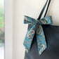 A Liberty Tana Lawn cotton skinny scarf featuring a teal blue paisley pattern, tied into a bow on a black handbag, shown hanging indoors next to a window.