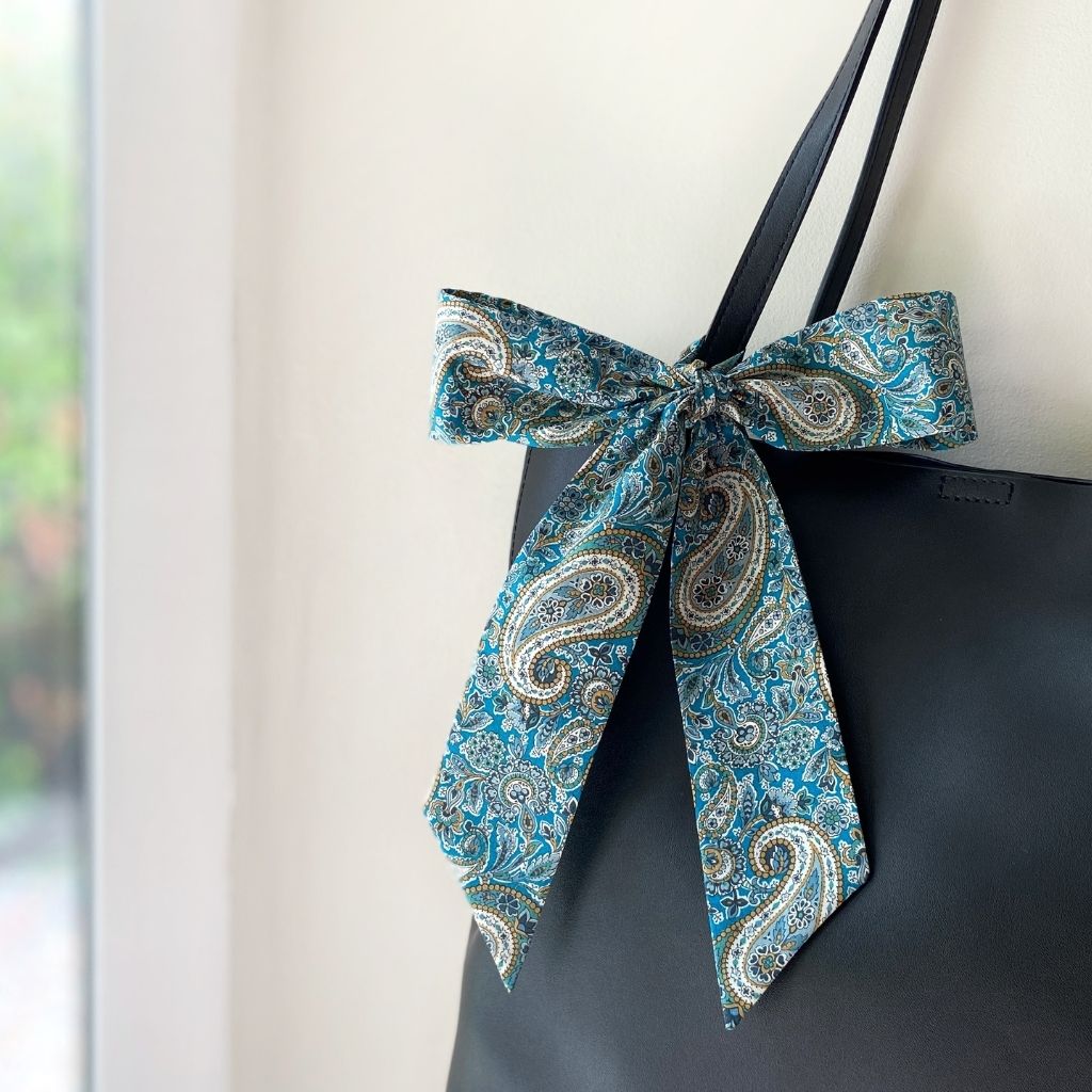 A Liberty Tana Lawn cotton skinny scarf featuring a teal blue paisley pattern, tied into a bow on a black handbag, shown hanging indoors next to a window.