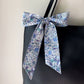A Liberty Tana Lawn cotton skinny scarf featuring a light blue floral pattern, tied into a bow on a black handbag, shown hanging against a gray wall.