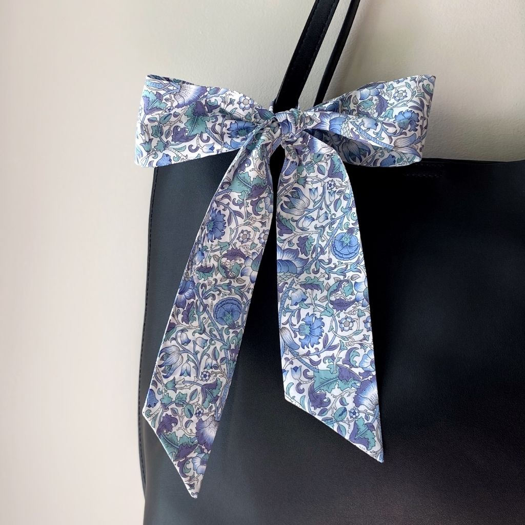 A Liberty Tana Lawn cotton skinny scarf featuring a light blue floral pattern, tied into a bow on a black handbag, shown hanging against a gray wall.