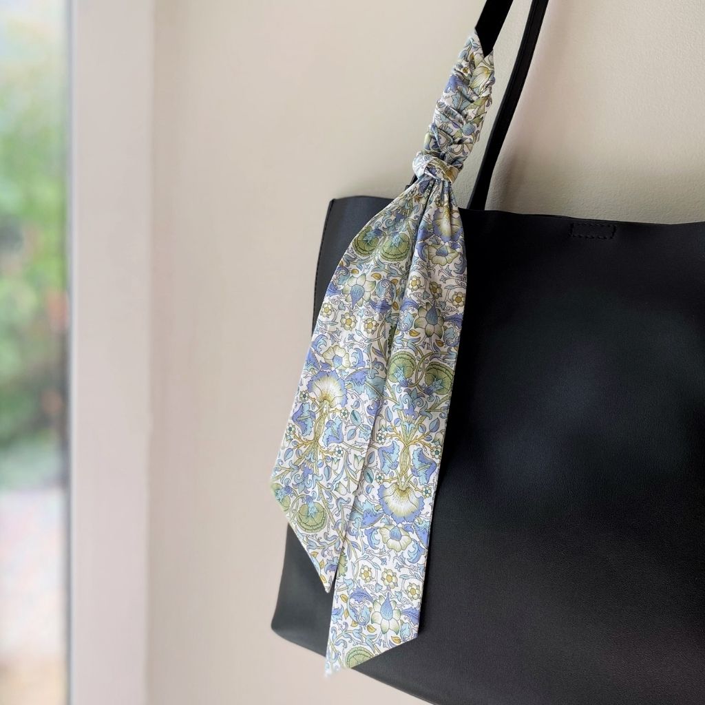 A Liberty Tana Lawn cotton skinny scarf featuring a light green, white, and blue floral pattern, tied on a black handbag, shown hanging indoors next to a window.