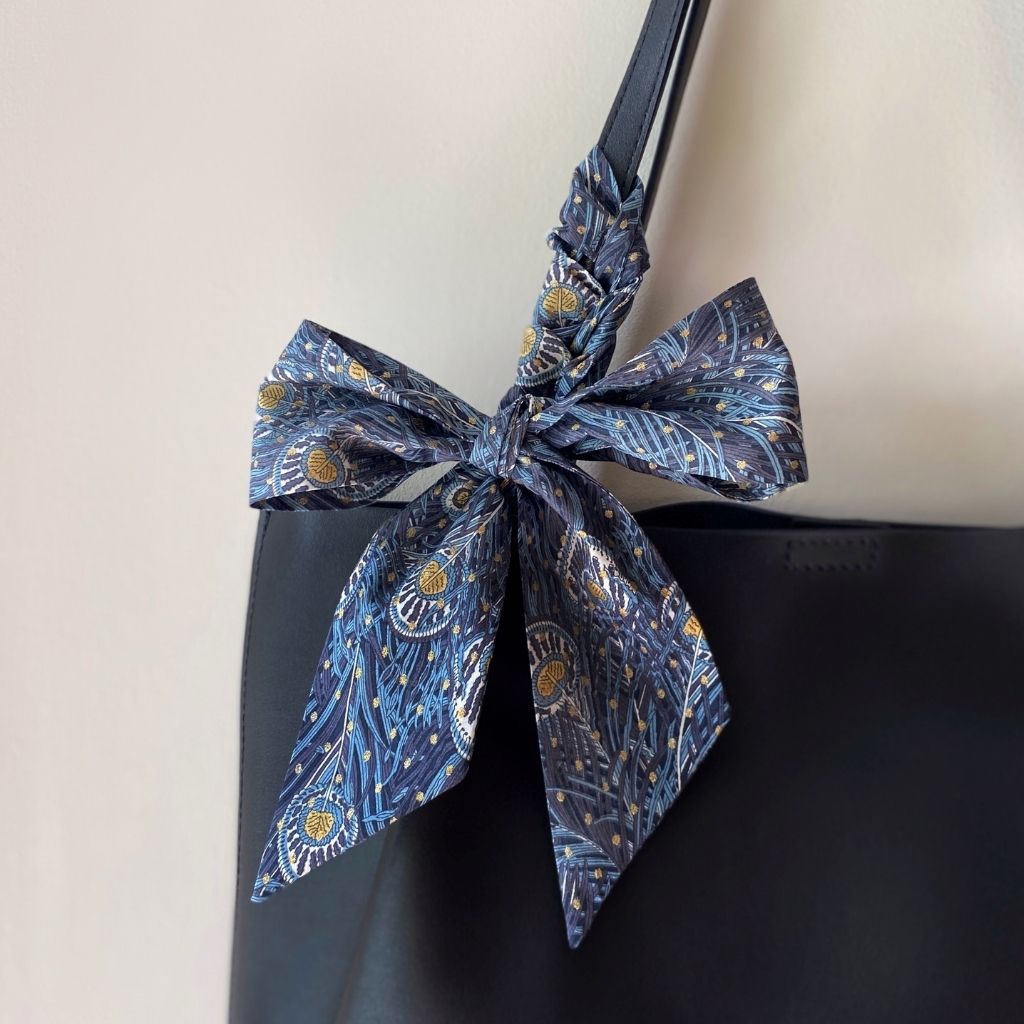 A gray-blue peacock feather patterned Liberty Tana Lawn cotton skinny scarf tied in a large bow and wrapped around the shoulder strap of a black tote bag, shown against a plain light background.