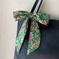 A green floral print Liberty Tana Lawn cotton skinny scarf tied into a large bow on a black tote bag's strap, against a plain light beige wall.