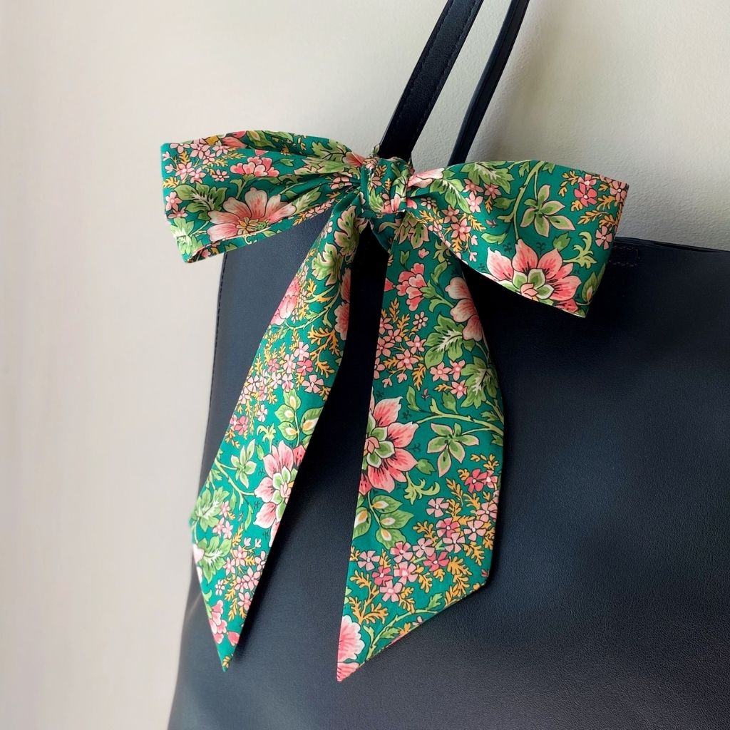 A green floral print Liberty Tana Lawn cotton skinny scarf tied into a large bow on a black tote bag's strap, against a plain light beige wall.