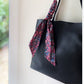 A black leather tote bag with a long Liberty Tana Lawn cotton skinny scarf tied to its strap, shown against a softly blurred indoor background.