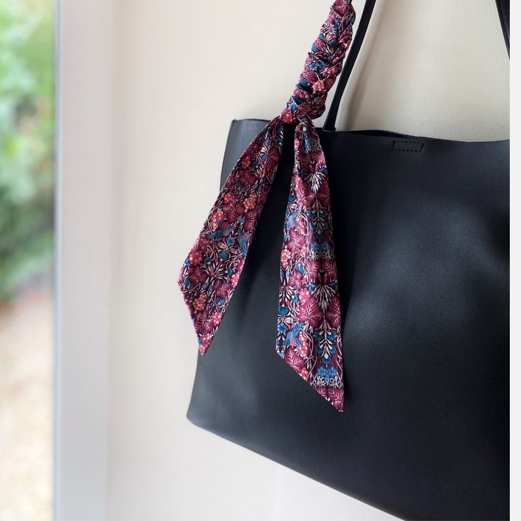 A black leather tote bag with a long Liberty Tana Lawn cotton skinny scarf tied to its strap, shown against a softly blurred indoor background.