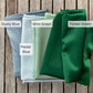 Four folded mulberry silk fabric swatches labeled 'Dusty Blue', 'Mint Green', 'Pastel Blue', and 'Forest Green' on a wooden surface.
