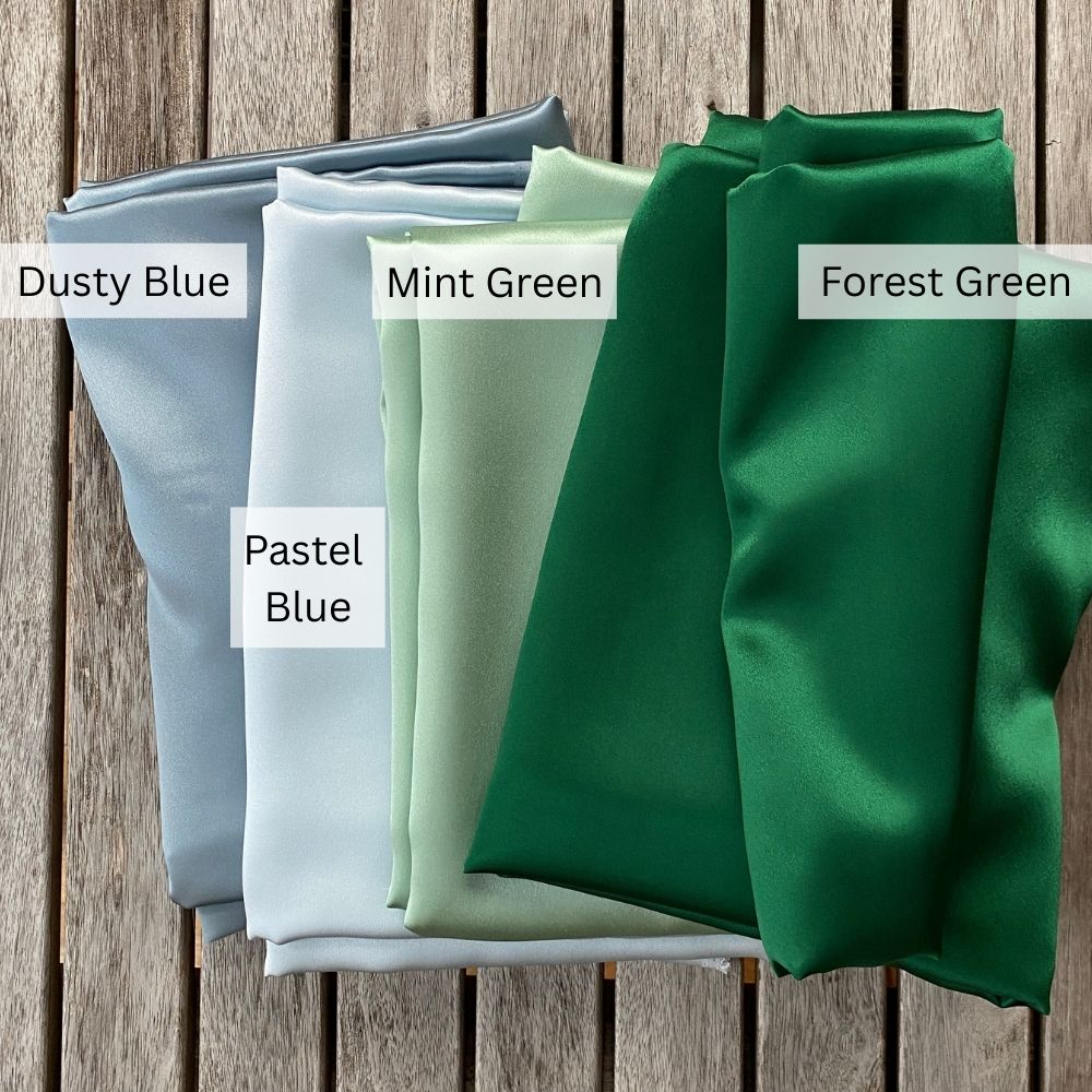Four folded mulberry silk fabric swatches labeled 'Dusty Blue', 'Mint Green', 'Pastel Blue', and 'Forest Green' on a wooden surface.
