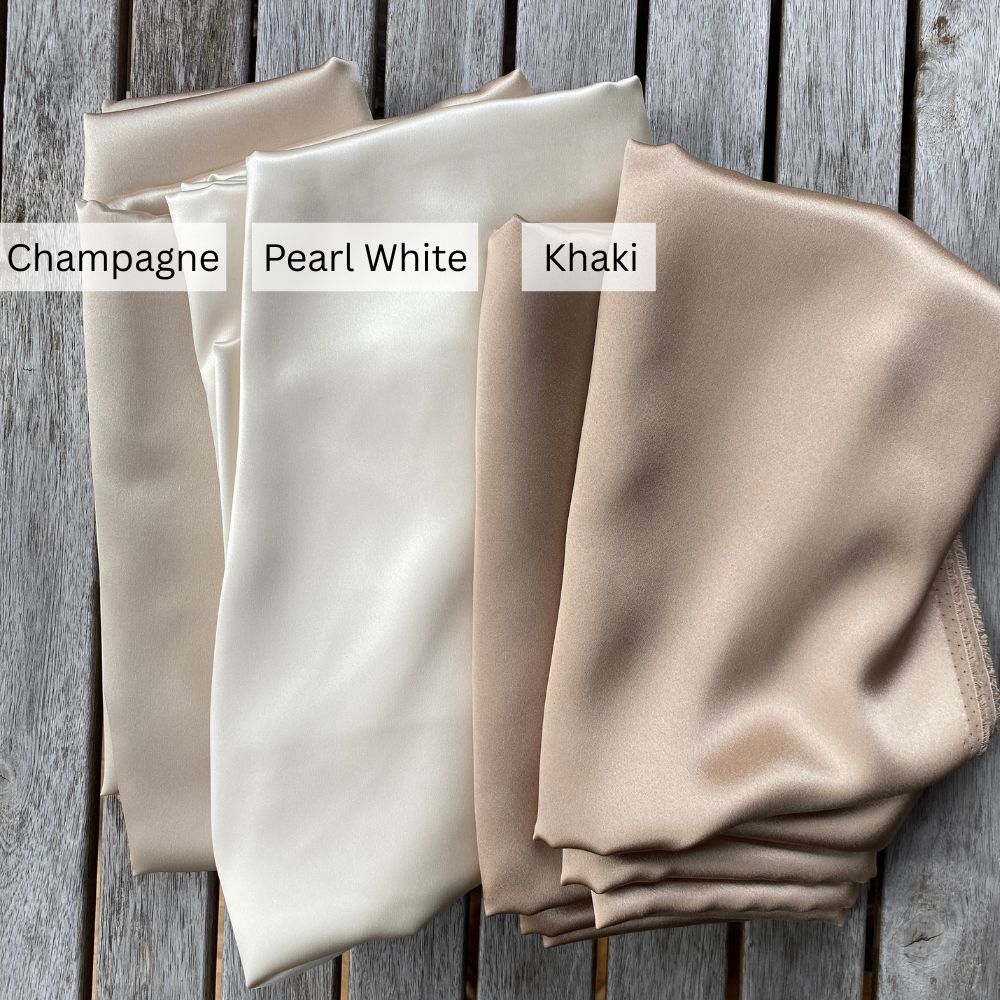Three folded mulberry silk satin fabric samples labeled 'Champagne', 'Pearl White', and 'Khaki' on a wooden surface.
