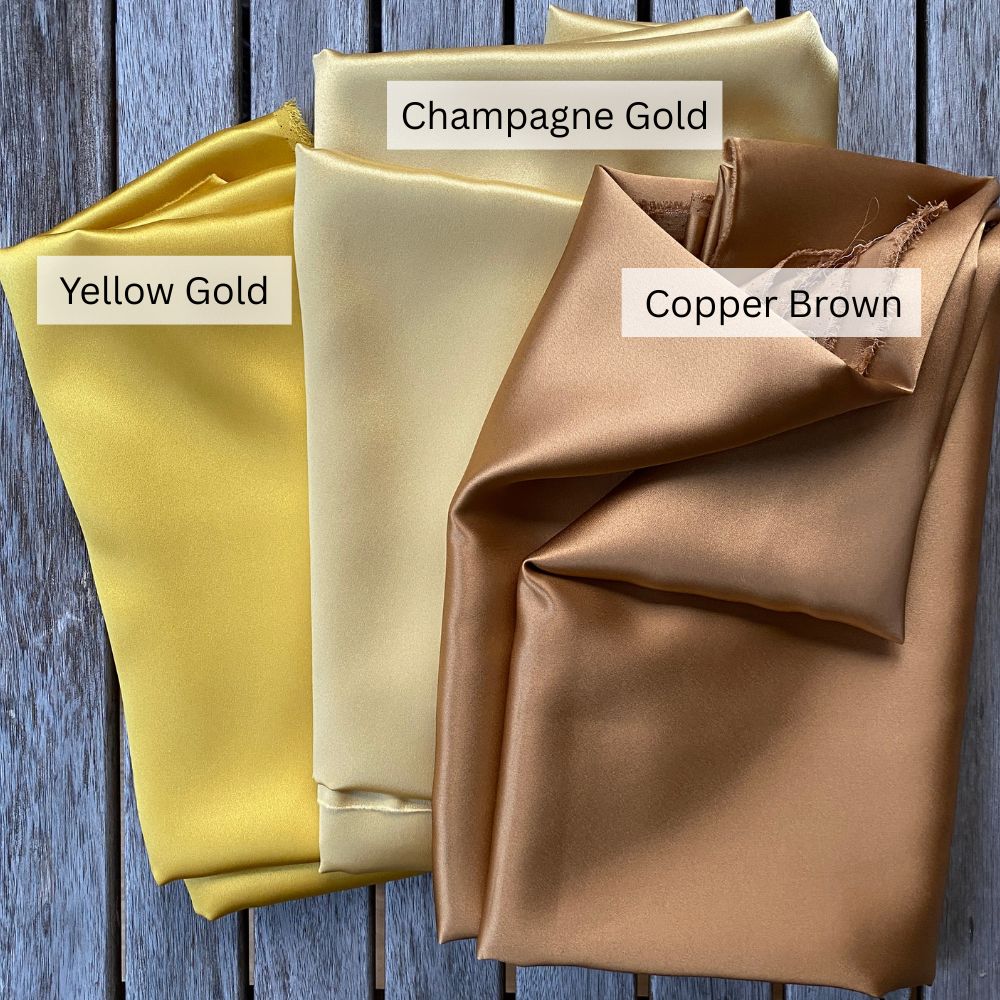 Three folded mulberry silk fabric swatches labeled 'Yellow Gold', 'Champagne Gold', and 'Copper Brown' on a wooden background.